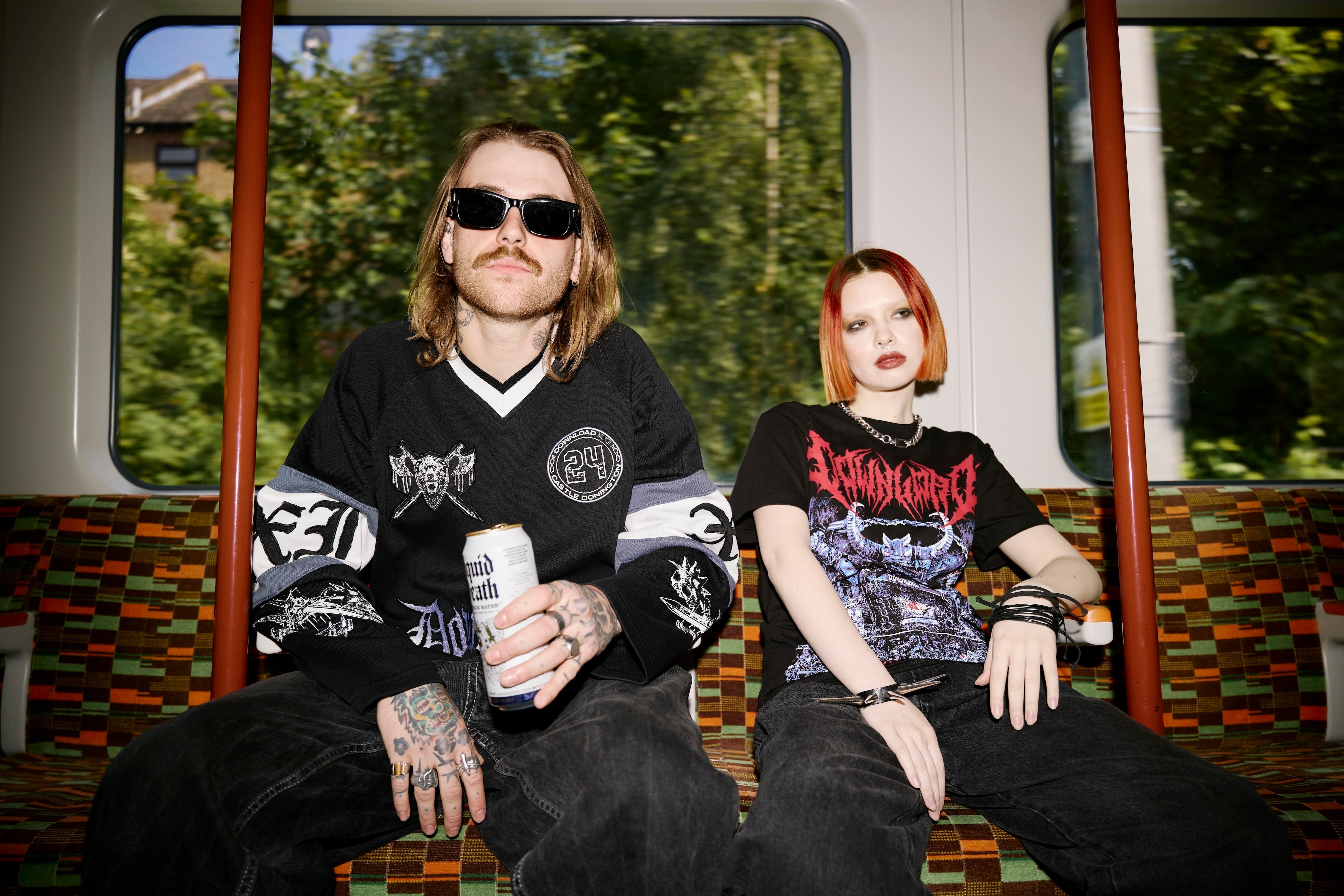 Male and female models sit on train carriage wearing MW DLXXI collection, male model is holding a can of liquid death water